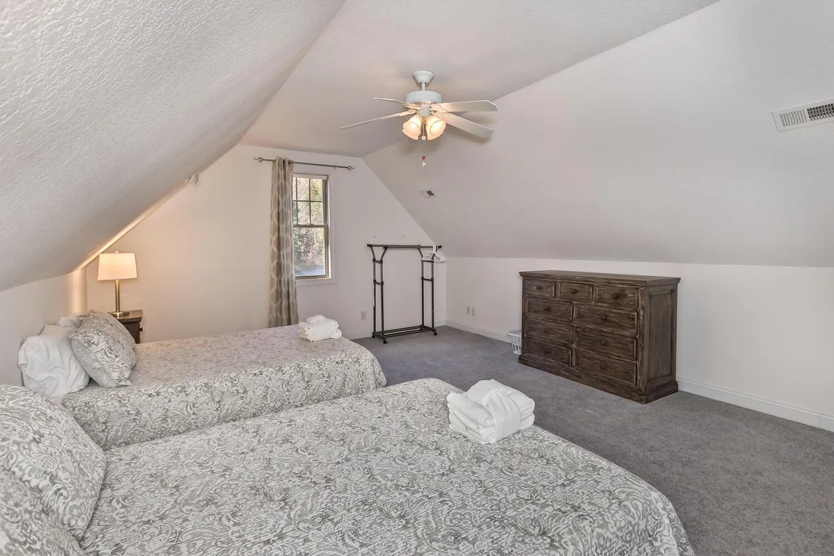 A comfortable bedroom with two twin beds, a dresser, window with curtains, and a ceiling fan. The space is set up for shared accommodation, offering an inviting atmosphere for teens, kids, or extra guests. Soft lighting and cozy bedding enhance the welcoming mood.