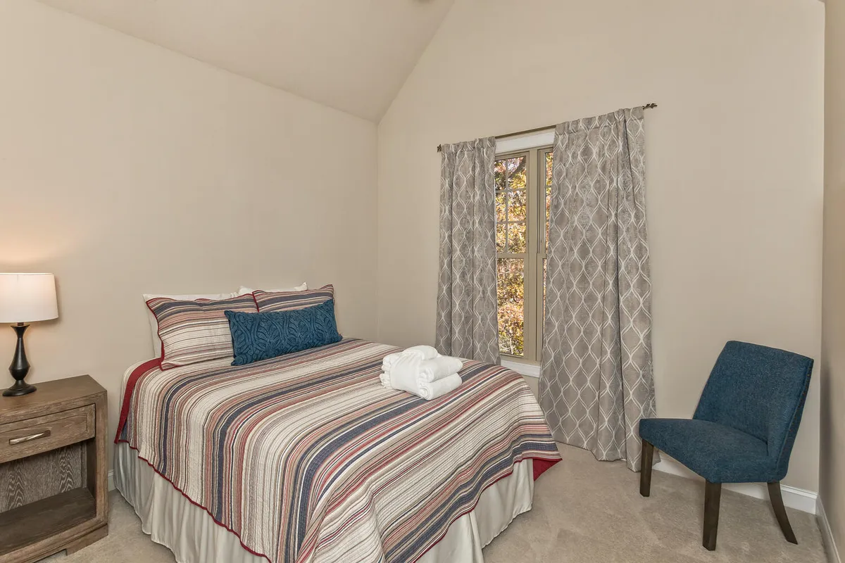 A restful bedroom with a bed dressed in striped bedding, a window with curtains allowing natural light, a nightstand with a lamp, and a blue chair in the corner. The scene feels inviting and restful, ideal for reading or enjoying a quiet morning.
