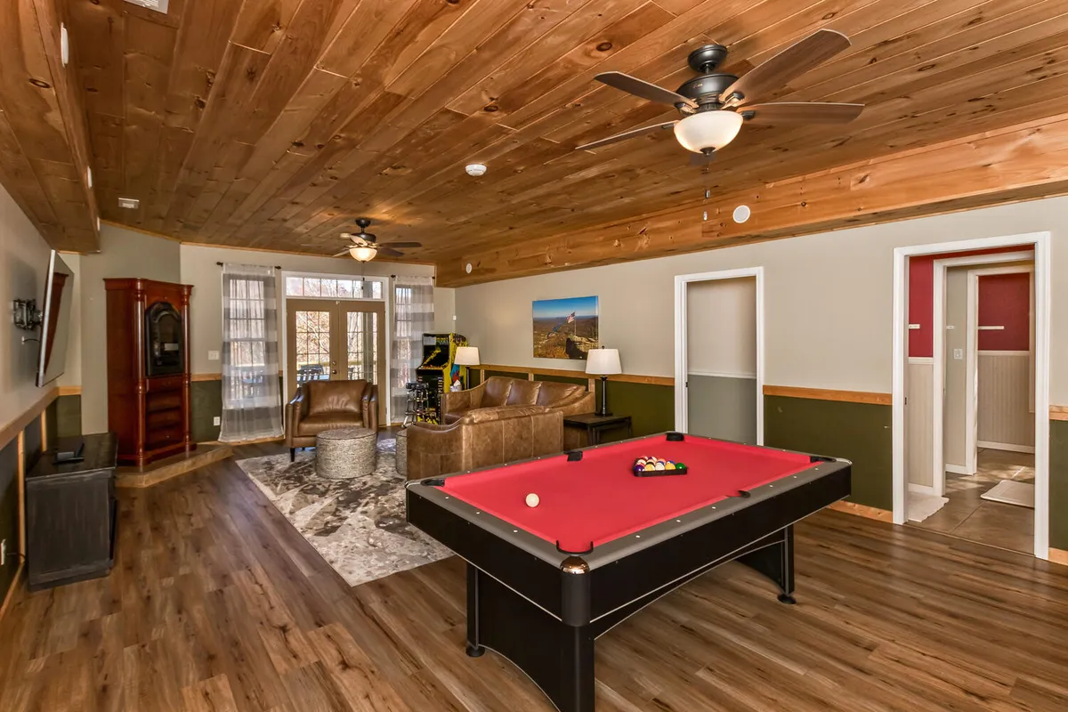 A spacious game and family room featuring a pool table at the center, surrounded by a comfortable seating area, rustic wooden ceiling, and large windows that let in plenty of natural light. A decorative rug adds warmth and the setup invites friendly competition or relaxed gatherings.