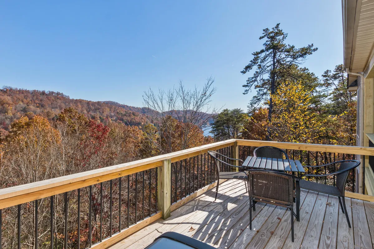 A peaceful exterior deck scene with black chairs and a table, set against autumn trees and a stunning lake view. The setup invites outdoor dining and relaxation during sunset or morning coffee.