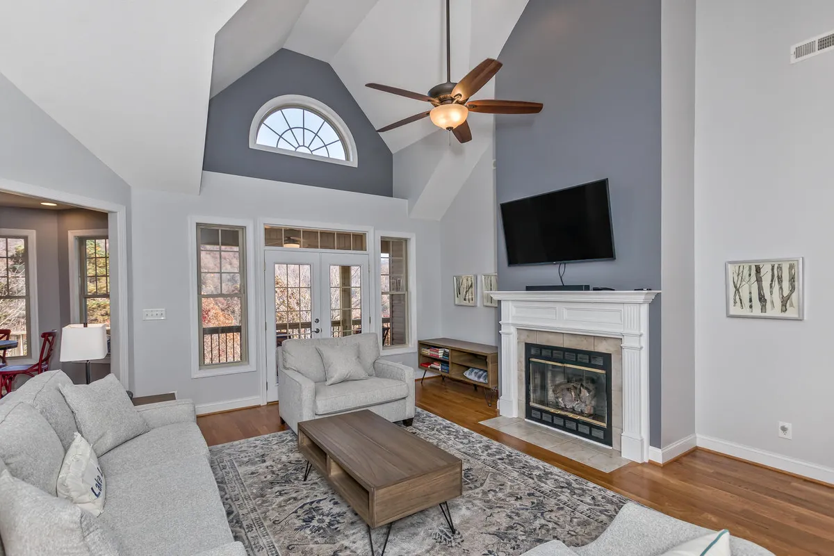 A cozy living room scene with a sofa set, a crackling fireplace, large windows revealing the outdoors, and a ceiling fan overhead. The space is styled with modern touches and offers a welcoming atmosphere ideal for relaxing, socializing, or enjoying the view.