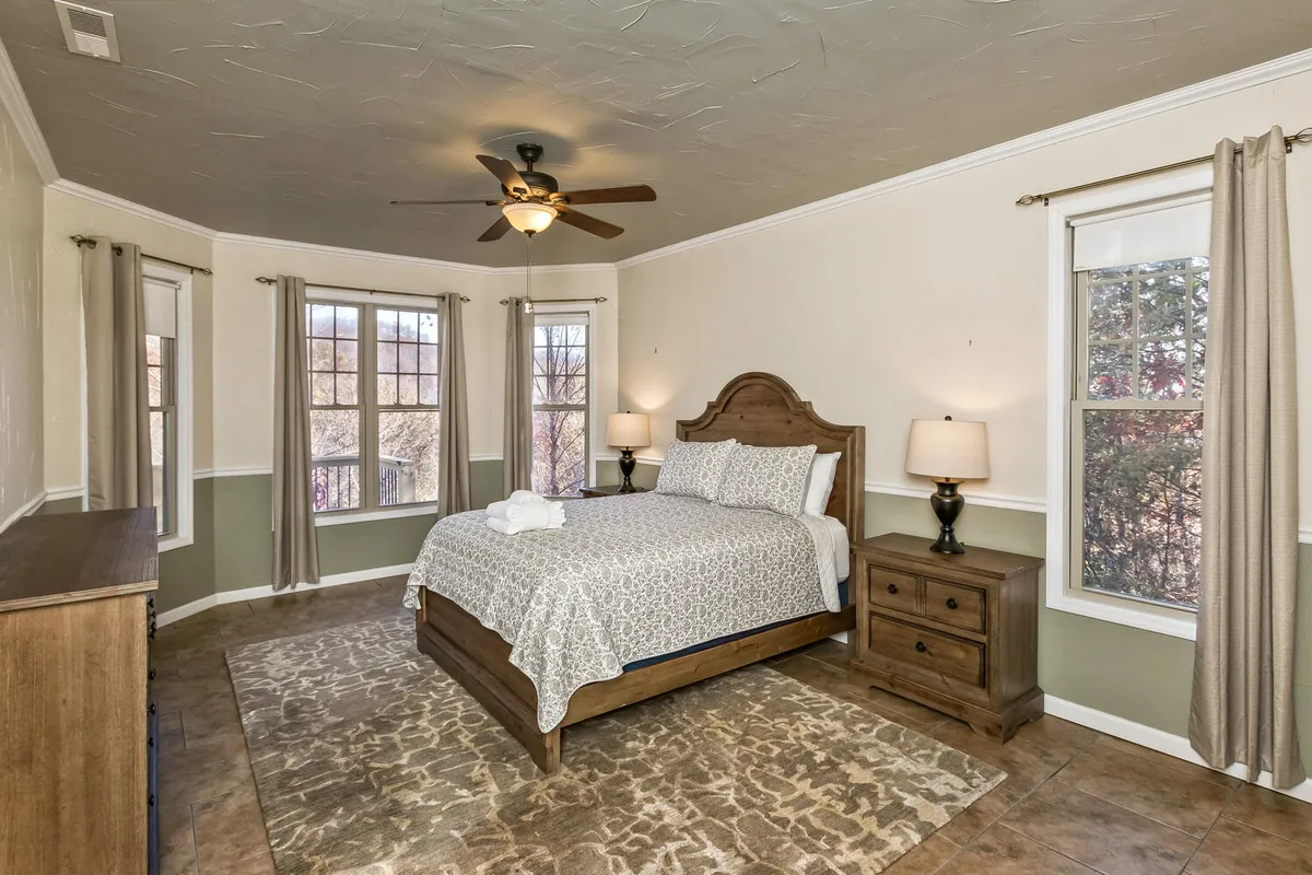 A spacious bedroom featuring a bed with patterned bedding, wooden dresser, bedside lamps, and large windows letting in abundant natural light. The setting feels inviting, with space for relaxation or reading by the window. The overall mood is cozy and tranquil, perfect for a restful retreat.