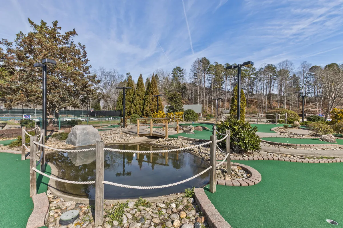 The image showcases a well-maintained miniature golf course with manicured lawns, a small pond crossed by a bridge, and attractive landscaping. Families and groups are seen enjoying a casual round of mini-golf, emphasizing friendly competition and togetherness.